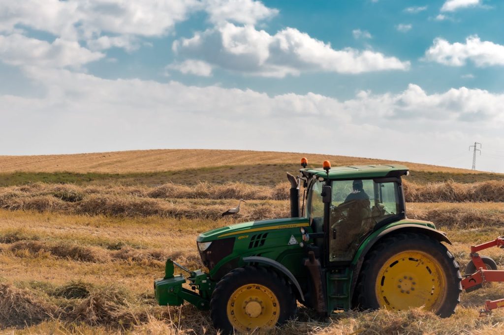 A green tractor plows through a rural field under a bright blue sky with clouds.